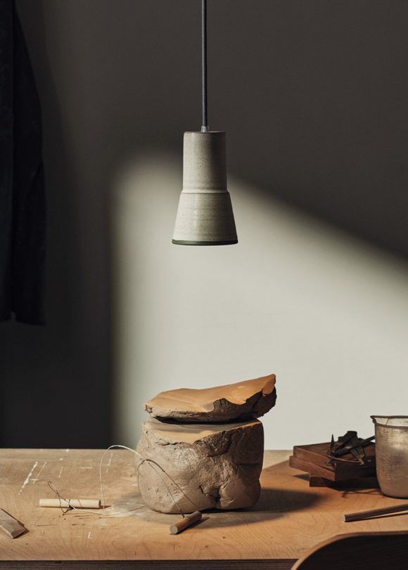 A stoneware pendant light, with a soft off-white glaze and iron speckles, hanging over a workspace with tools and materials visible.